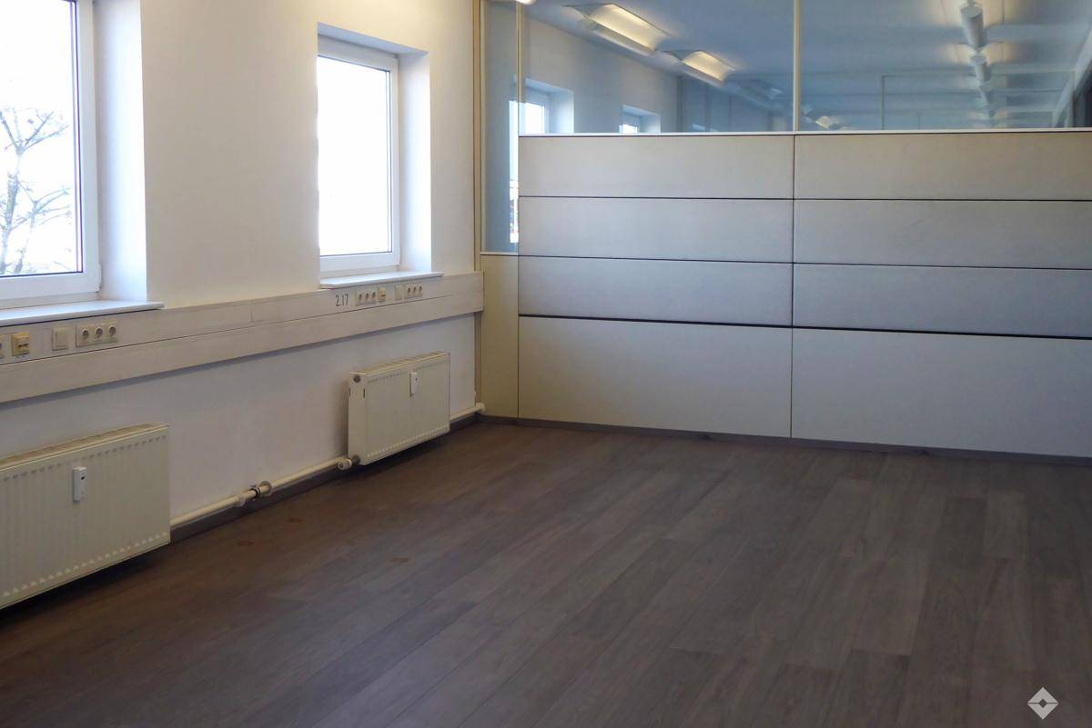 Leerer Büroraum mit weißen Wänden, großen Fenstern und Heizkörpern, ideal für Arbeitsplätze.