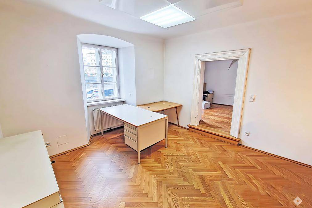Geräumiges Büro mit klassischem Fischgrätparkett und einfacher Büroeinrichtung, gut beleuchtet.