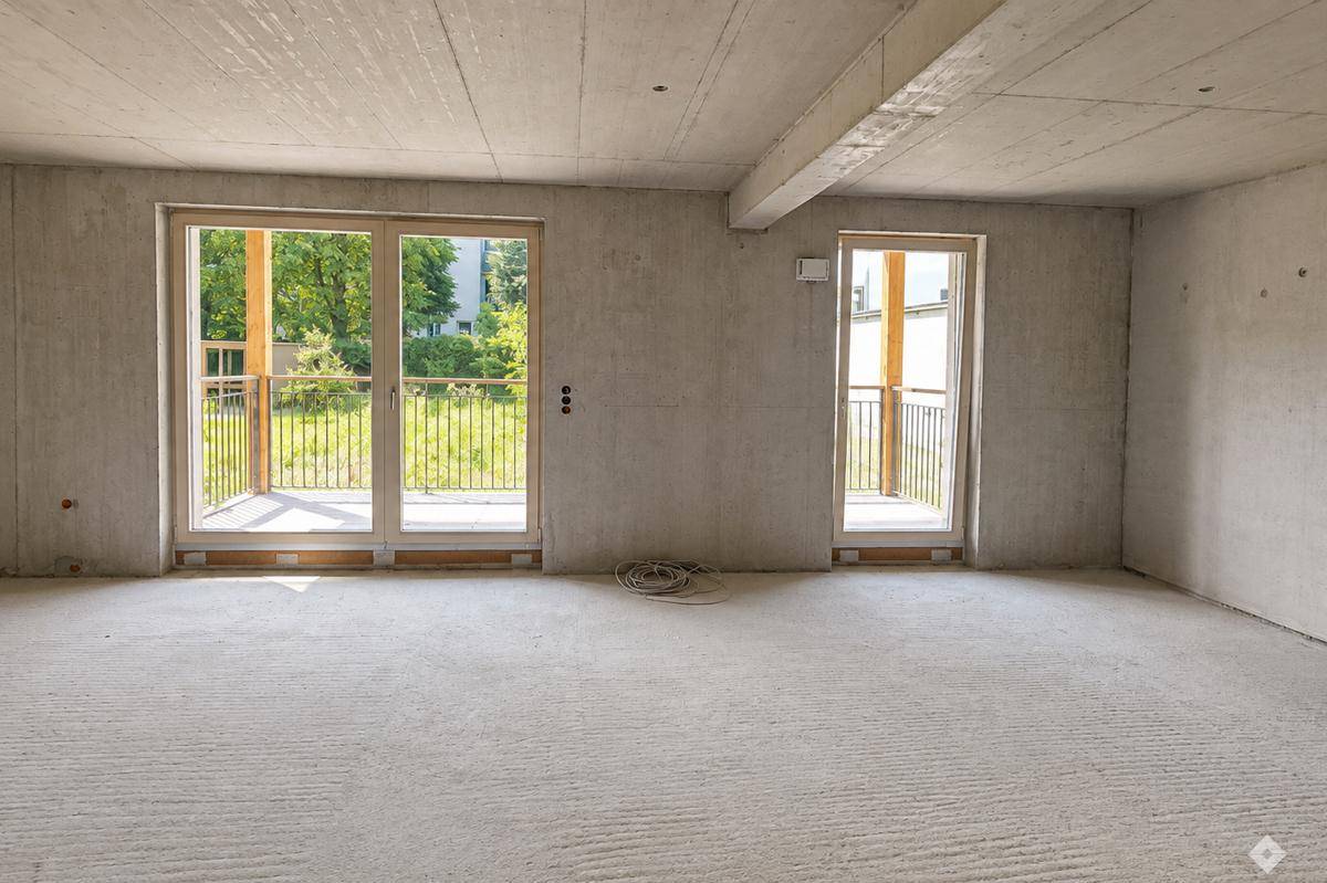 Unfertiger Innenraum mit großen Fenstern und Zugang zu einem Balkon, der viel natürliches Licht hereinlässt.