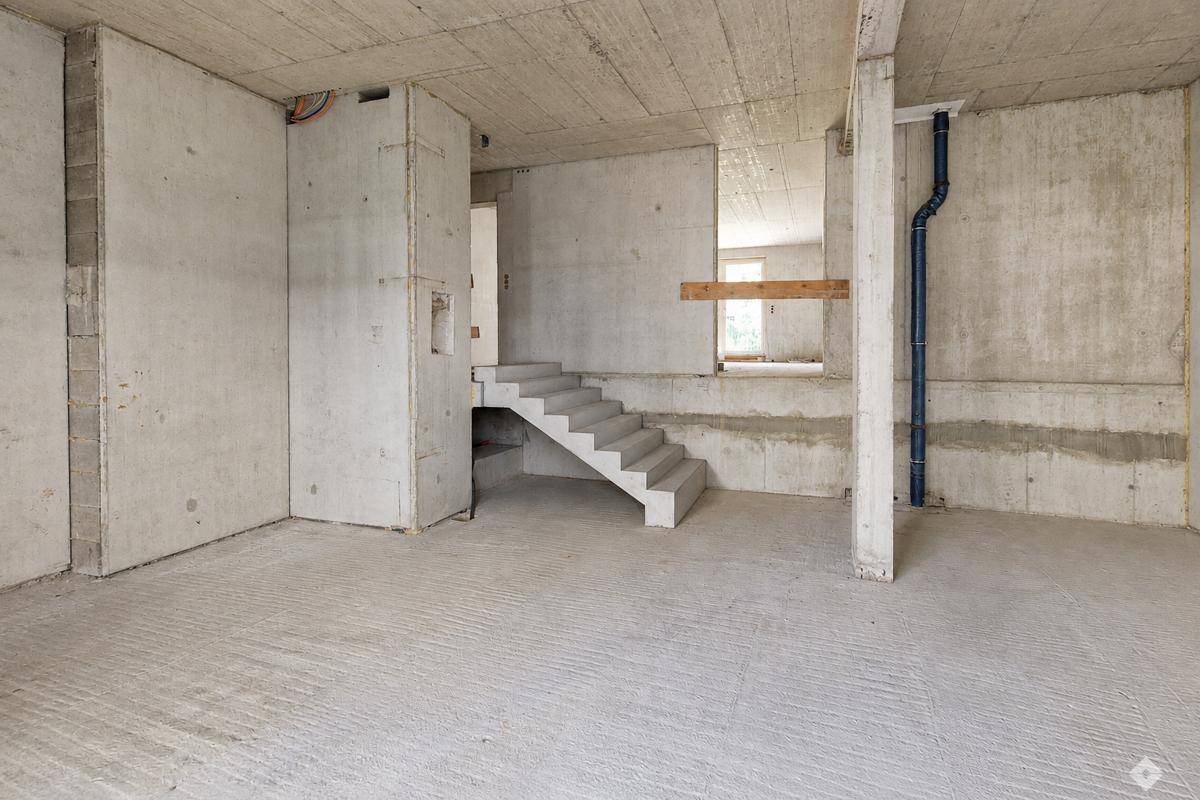 Rohbau-Innenraum mit freiliegenden Betonwänden, einem Treppenaufgang und sichtbaren Installationen.