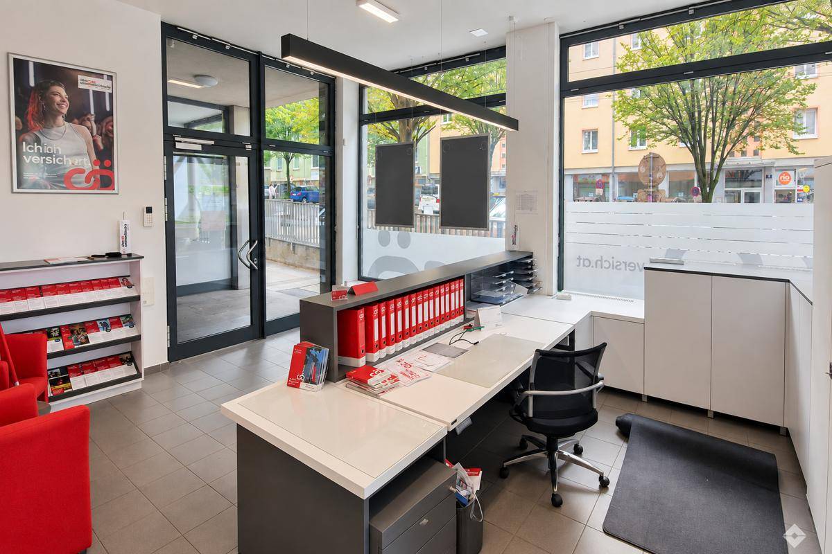 Empfangsbereich eines Büros mit Schreibtisch, roten Akzenten und großen Fenstern zur Straße.