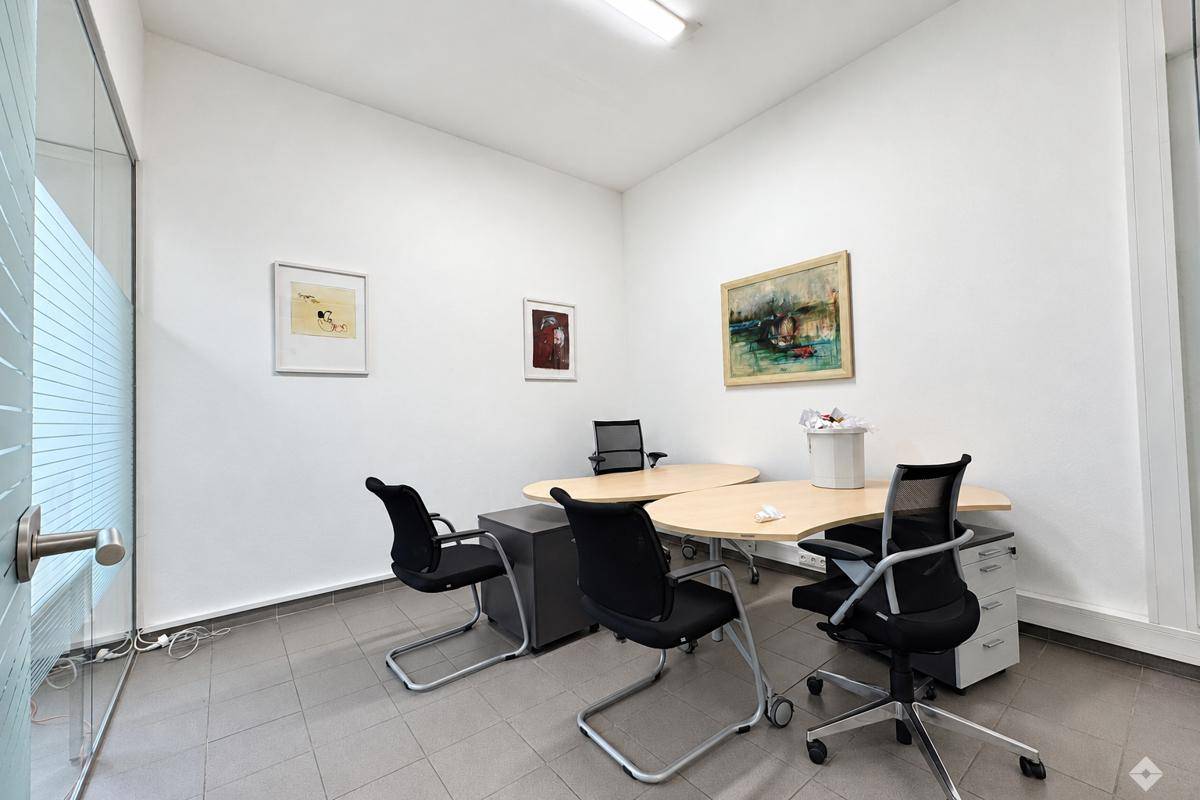 Kleineres, helles Büro mit weißen Wänden, Fliesenboden und zwei Schreibtischen.