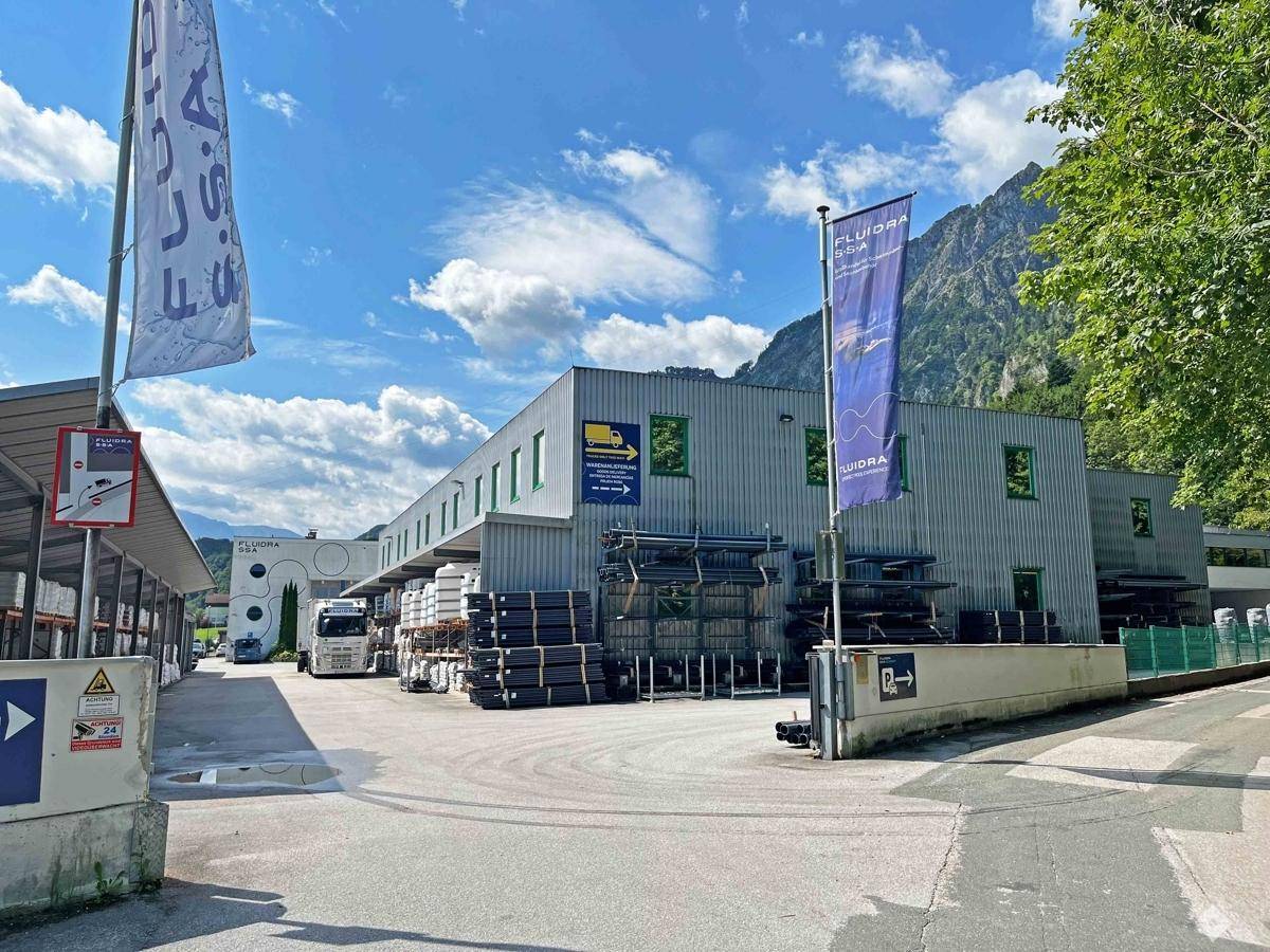 Industriegebäude mit Lagerflächen und Büros, umgeben von Bergen und Bäumen unter blauem Himmel.