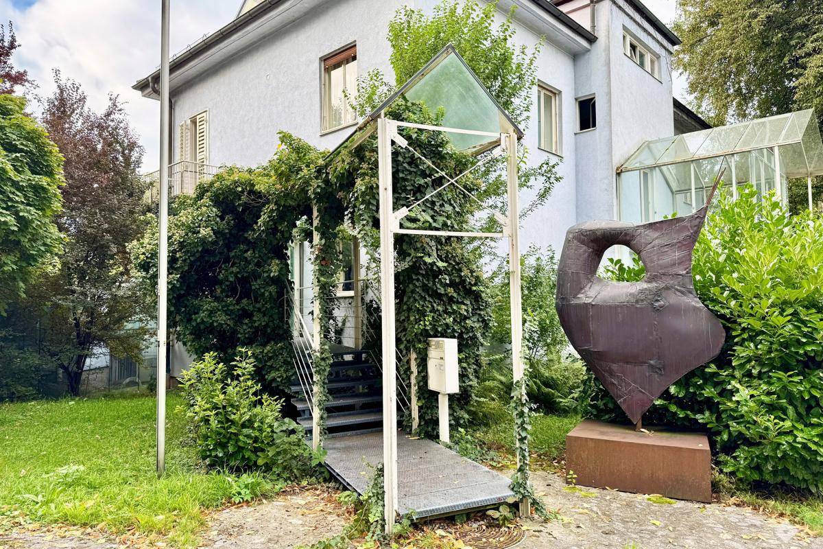 Außenansicht eines Gebäudes mit begrüntem Eingangsbereich und moderner Skulptur im Vorgarten.