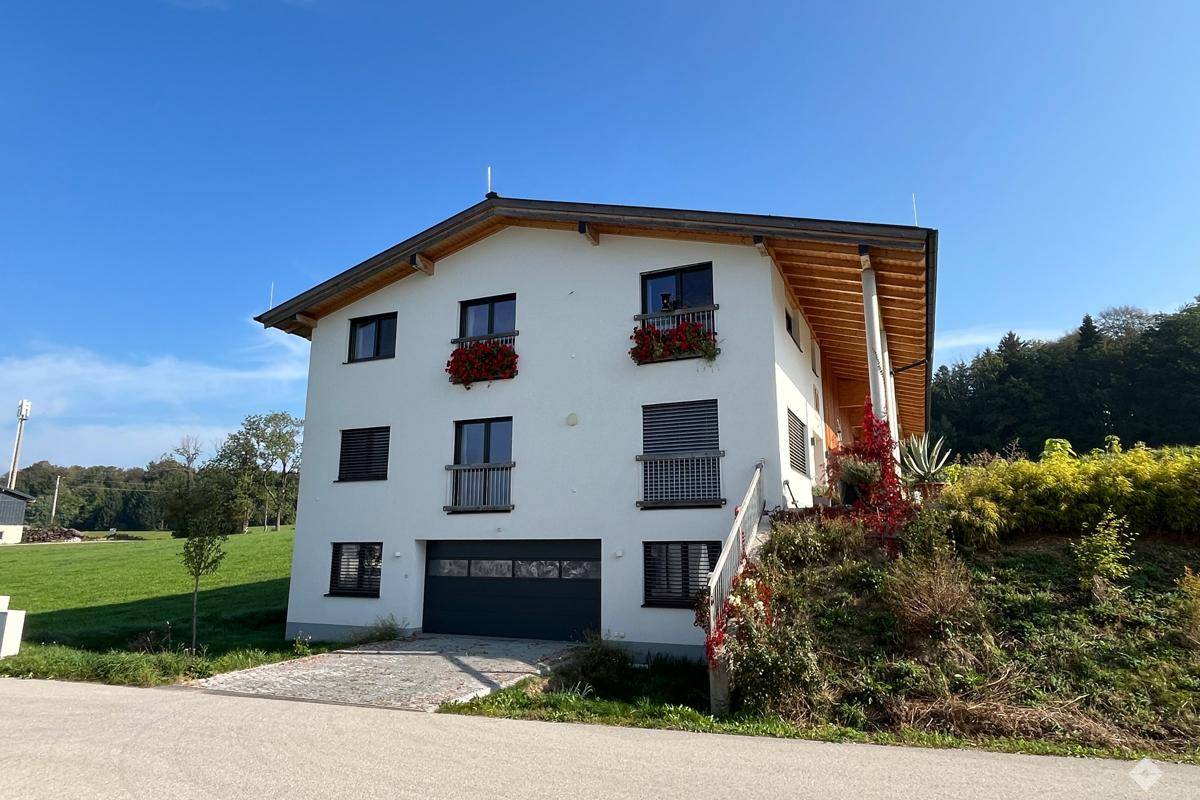 Vorderansicht des Gebäudes mit weißer Fassade, Balkonen und Garagentor, umgeben von Grünflächen.