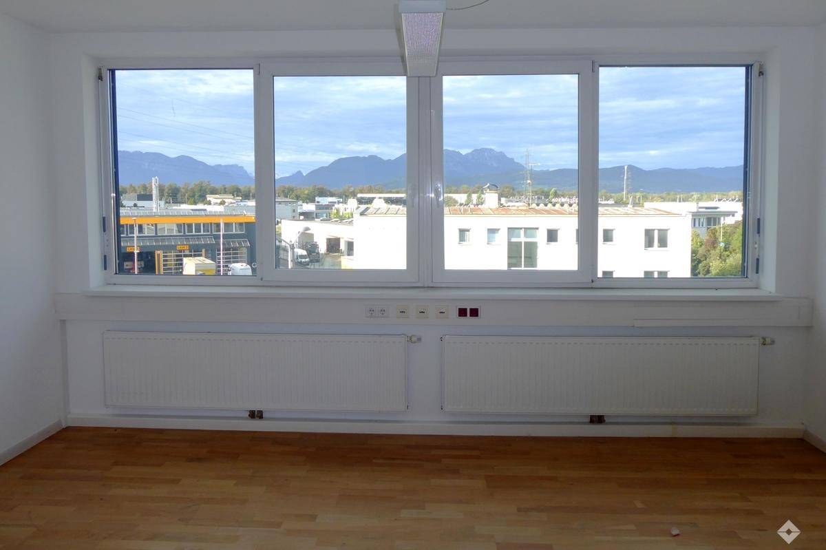 Große Fensterfront mit Blick auf eine städtische Umgebung und ferne Berge, in einem Raum mit Holzfußboden.