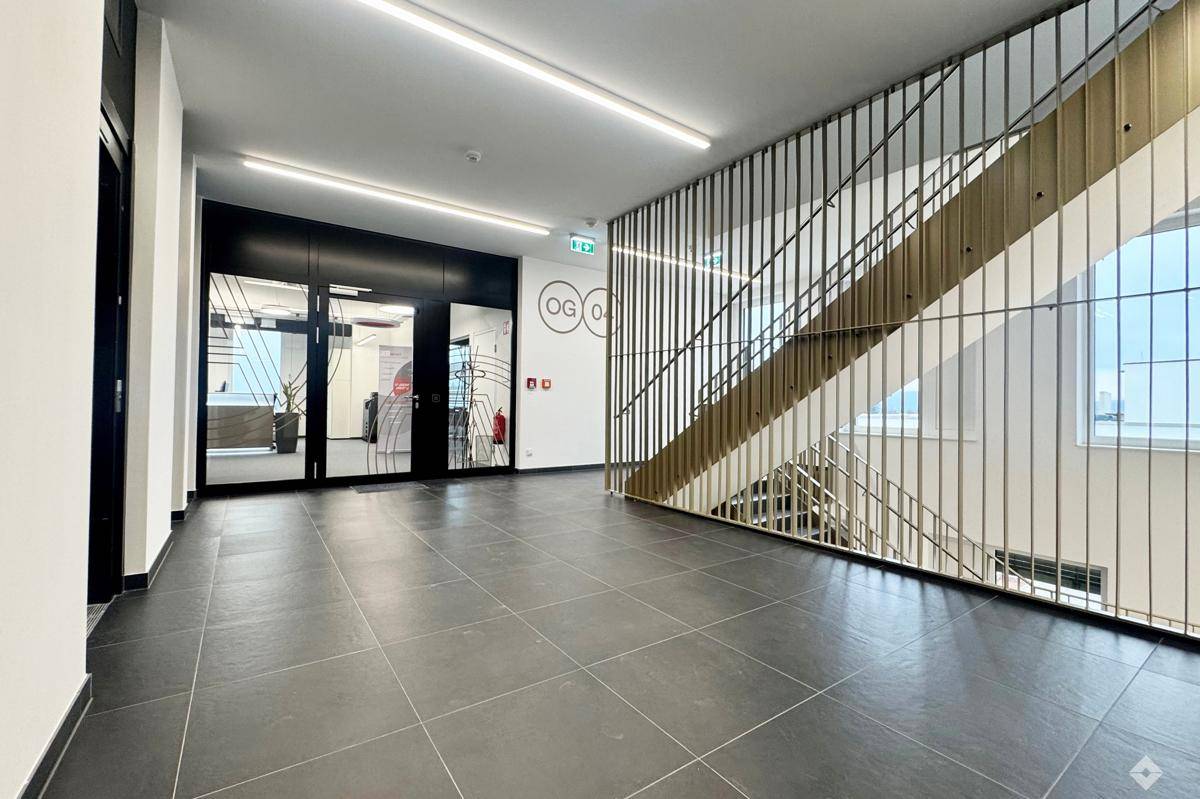 Heller Flur mit dunklen Fliesen, moderner Treppe und Glasfronten zu den Büroräumen.
