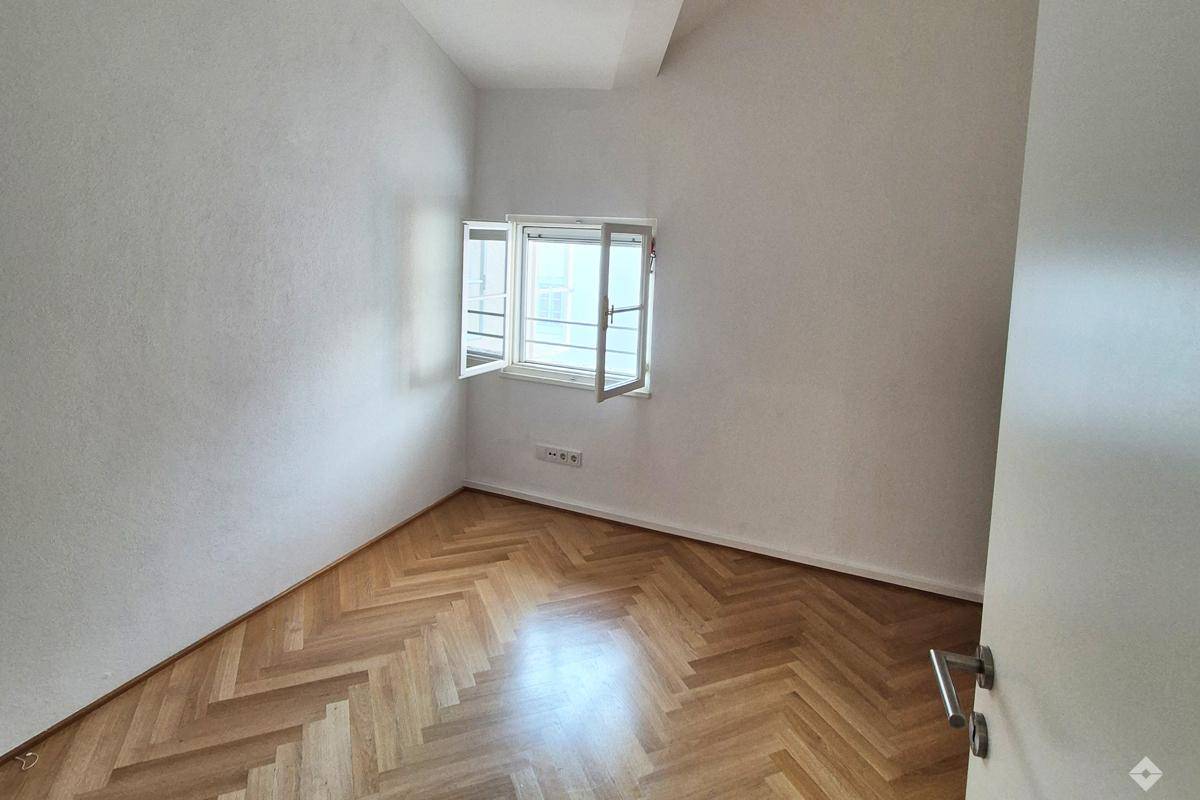 Kleines, helles Zimmer mit Fischgrätparkett und einem geöffneten Fenster, ideal als Schlaf- oder Arbeitszimmer.