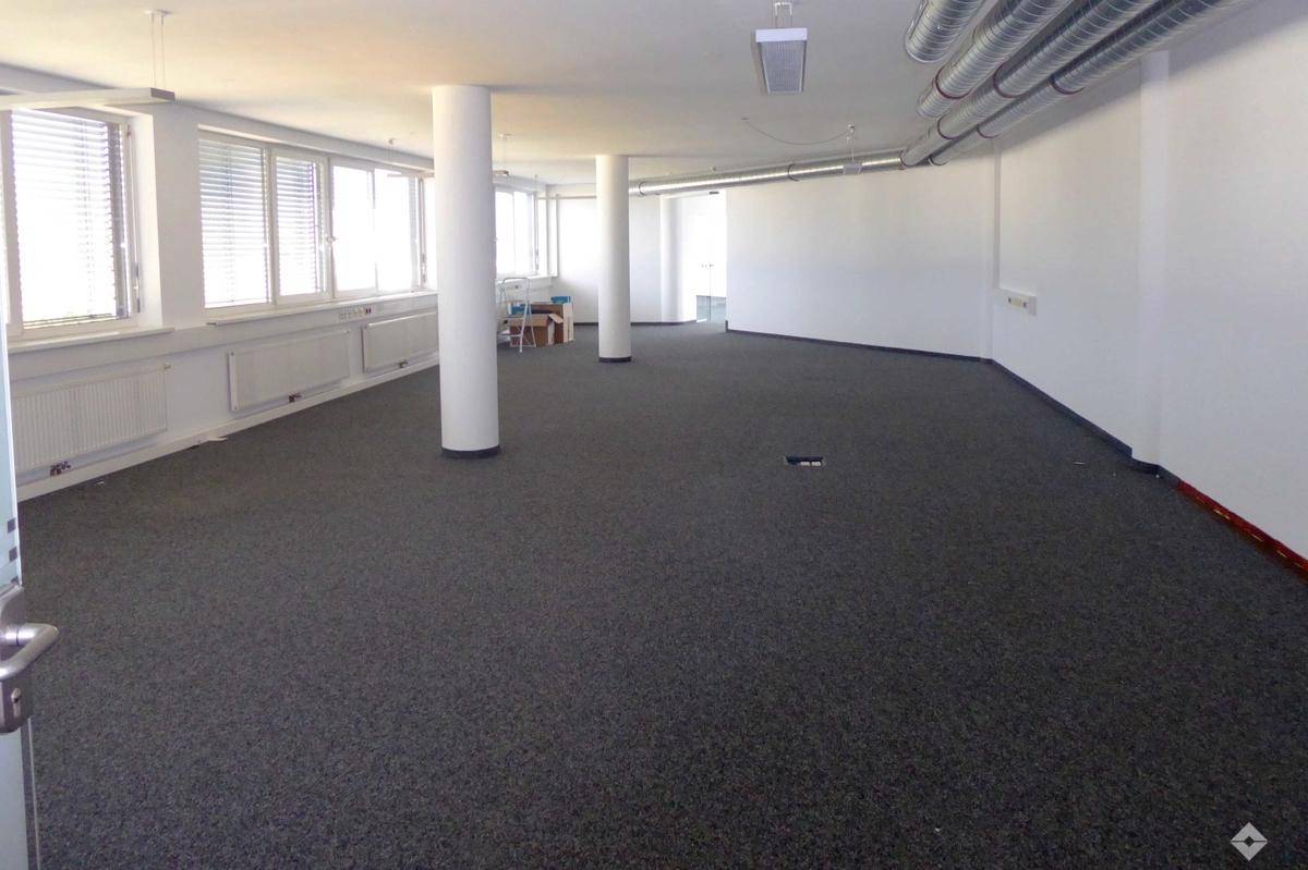 Großer, offener Büroraum mit Teppichboden, vielen Fenstern und sichtbaren Lüftungsrohren an der Decke.
