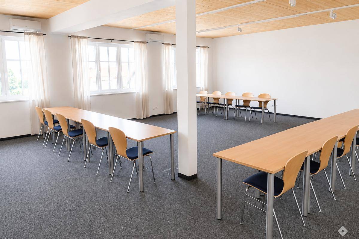 Großer, heller Konferenzraum mit Holzbalkendecke, Teppichboden und mehreren Tischen für Besprechungen.
