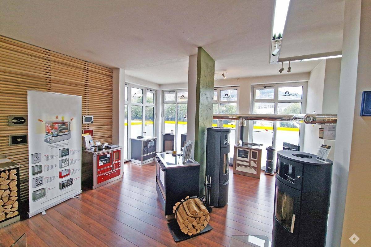 Heller Ausstellungsraum mit Holzboden, großen Fenstern und verschiedenen Kaminöfen als Exponate.