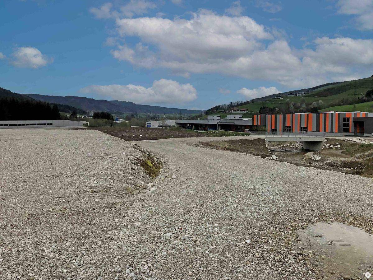 Unbebautes Kiesgrundstück mit Fahrweg, dahinter moderne Industriegebäude und grüne Landschaft unter blauem Himmel.