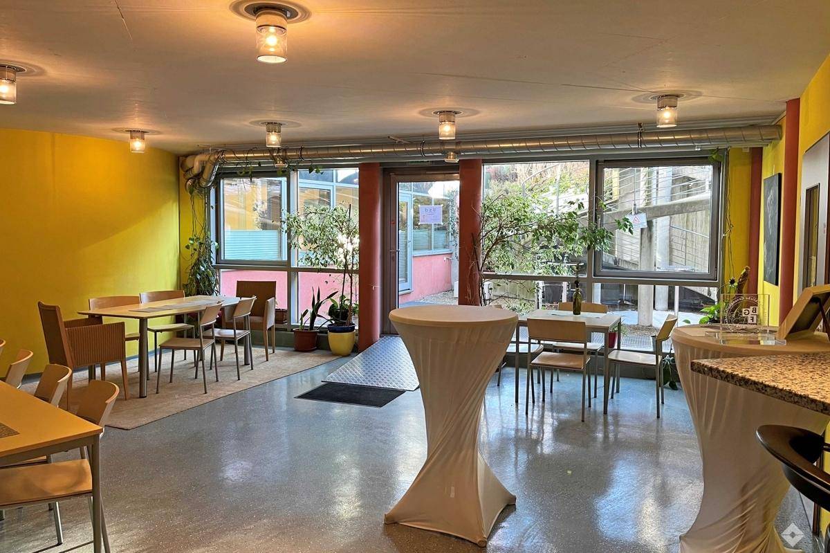 Vielseitiger Gemeinschaftsraum mit Tischen und Stehtischen, großen Fenstern und Blick auf einen Innenhof.