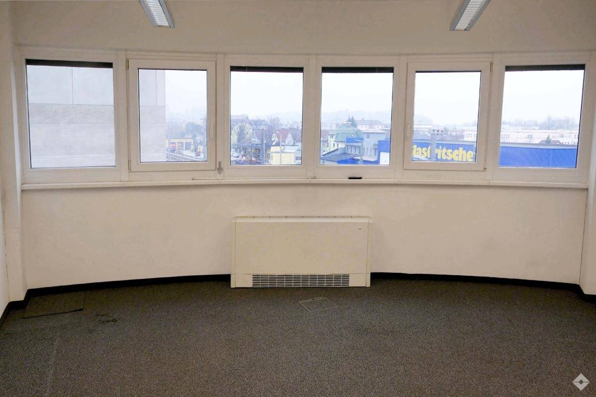 Büroraum mit Teppichboden und einer geschwungenen Fensterfront, die einen weiten Blick auf die städtische Umgebung bietet.