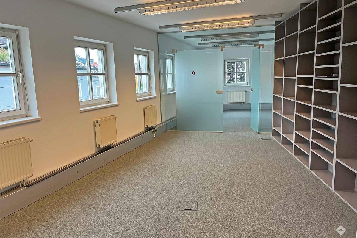 Geräumiger Büroraum mit Teppichboden, Einbauregalen und Fenstern, die viel Licht hereinlassen.