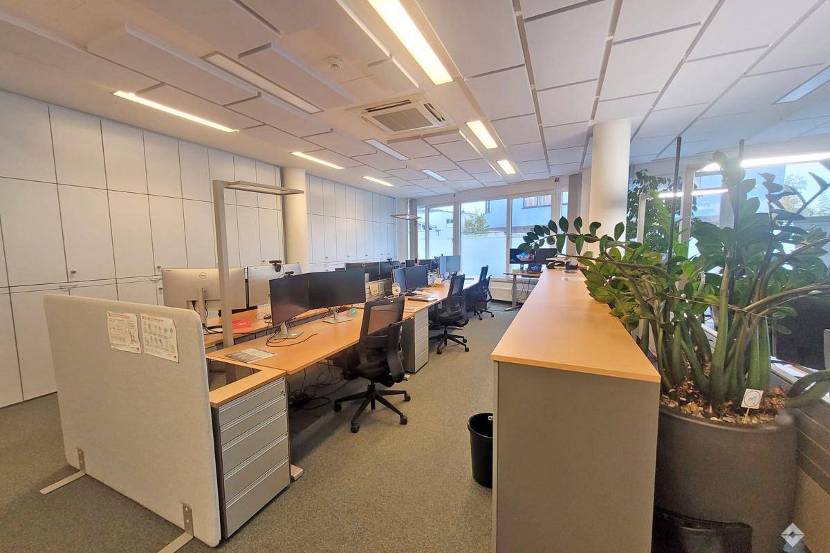 Modernes Großraumbüro mit mehreren Arbeitsplätzen, großen Fenstern, abgehängter Decke und Klimaanlage.