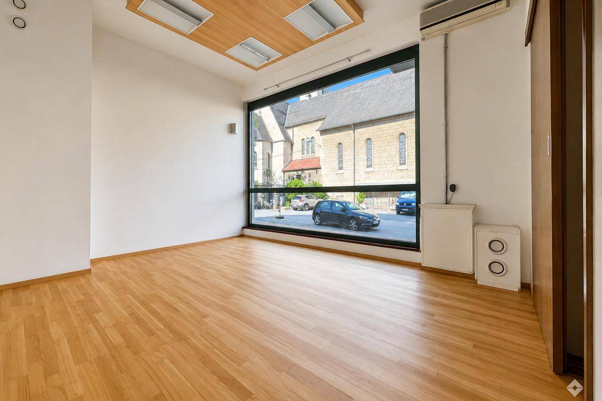 Leerer Raum mit hellem Holzfußboden, weißen Wänden und einem Fenster mit Jalousien.