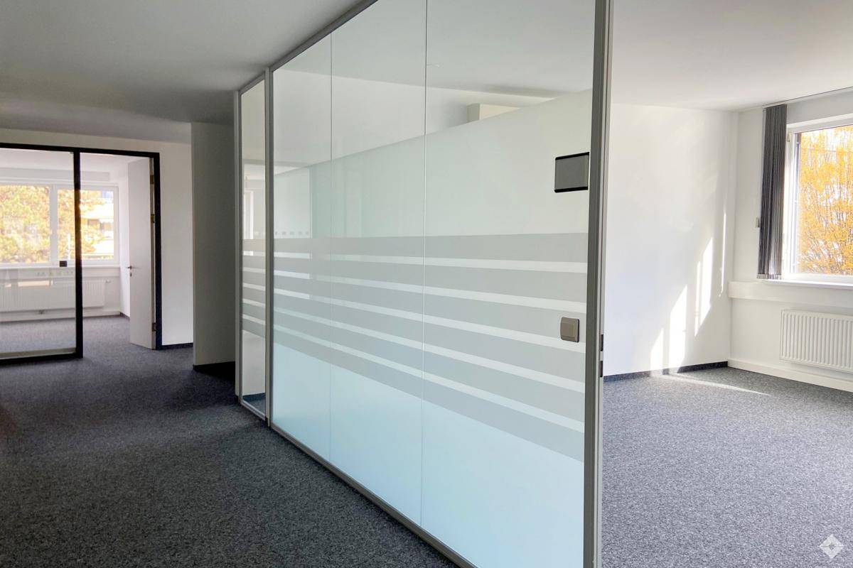 Moderner Bürobereich mit Glaswänden und Teppichboden, der verschiedene Arbeitsbereiche abtrennt.