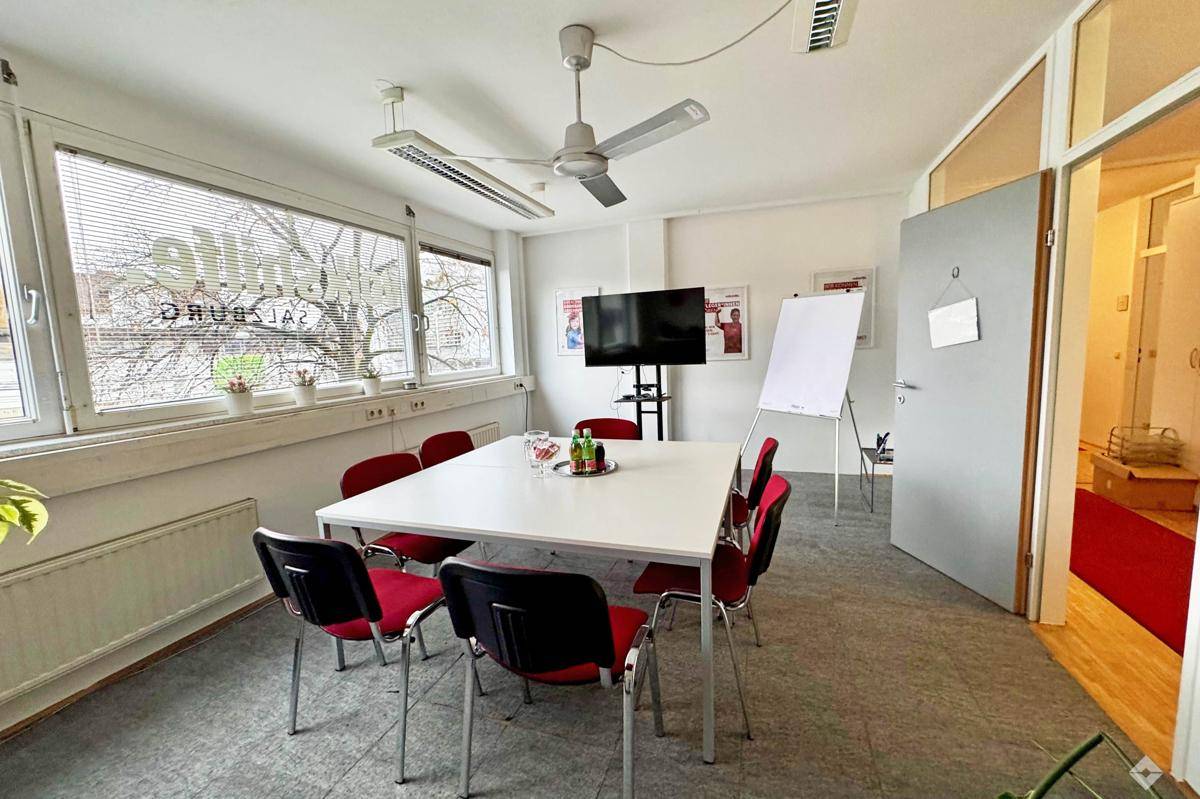 Konferenzraum mit großem Tisch, roten Stühlen, Deckenventilator und großen Fenstern.