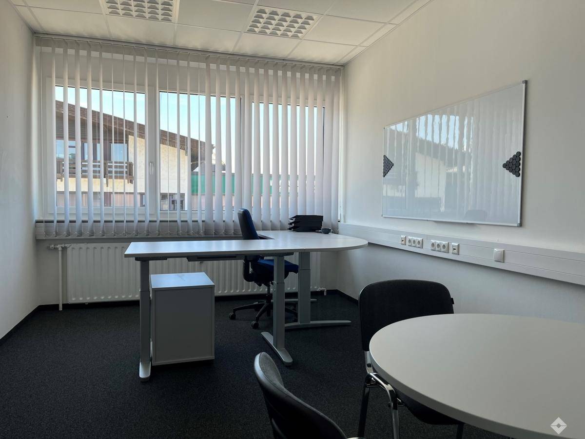 Geräumiges Büro mit Teppichboden, vertikalen Jalousien und einem großen Whiteboard an der Wand.