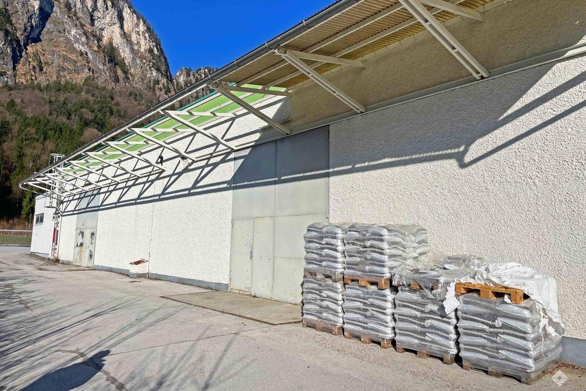 Seitenansicht des Industriegebäudes mit überdachtem Bereich und gestapelten Paletten vor einer Berglandschaft.