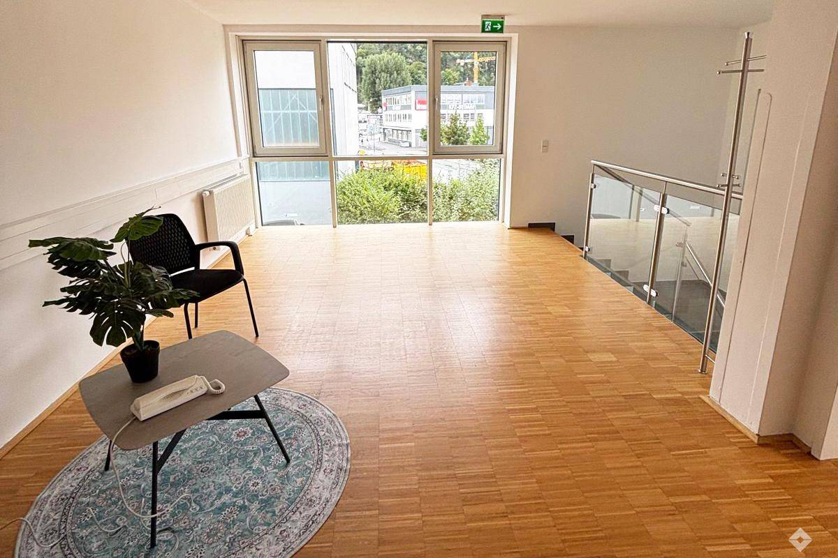 Heller Gemeinschaftsbereich mit großen Fenstern, Holzfußboden und einer modernen Treppe mit Glasgeländer.