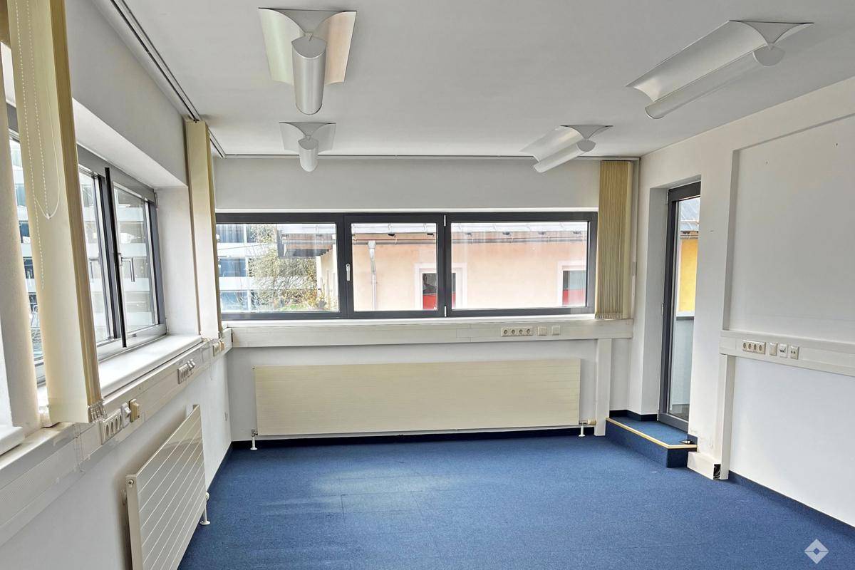 Funktionales Büro mit großen Fenstern, vertikalen Jalousien und Zugang zu einem weiteren Bereich.