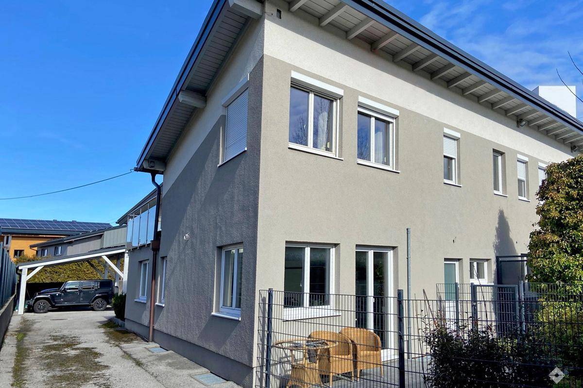 Modernes Gebäude mit heller Fassade, großen Fenstern und einem überdachten Carport auf der linken Seite.