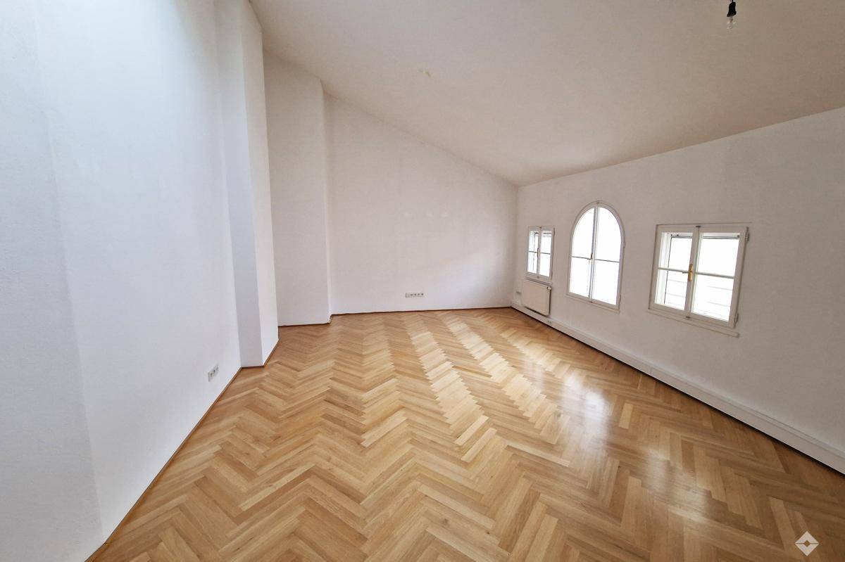 Heller Wohnbereich mit Fischgrätparkett, Dachschräge und mehreren Fenstern für viel Tageslicht.