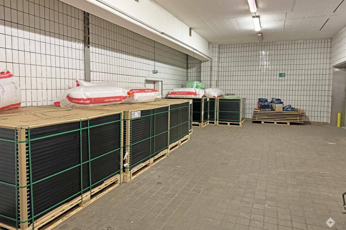 Lagerraum mit gestapelten Paletten voller verpackter Güter, gefliesten Wänden und Deckenbeleuchtung.