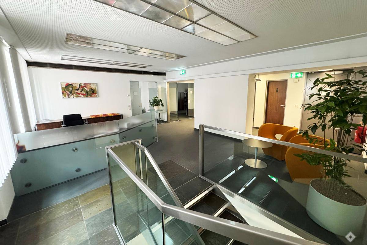Geräumiges Büro mit Teppichboden, Schreibtisch und Blick auf die Treppe, gut beleuchtet durch ein Fenster.