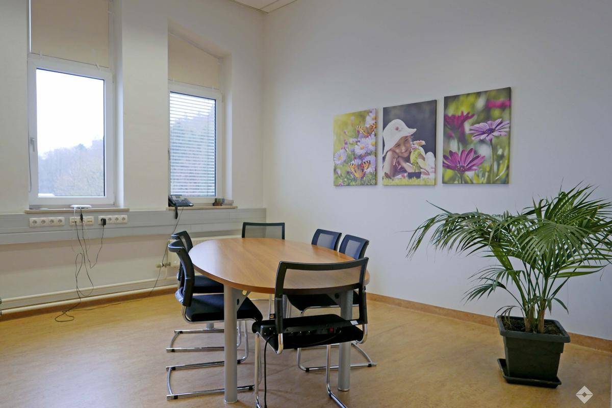 Besprechungsraum mit ovalem Tisch, Stühlen, Wandbildern und einer Pflanze am Fenster.