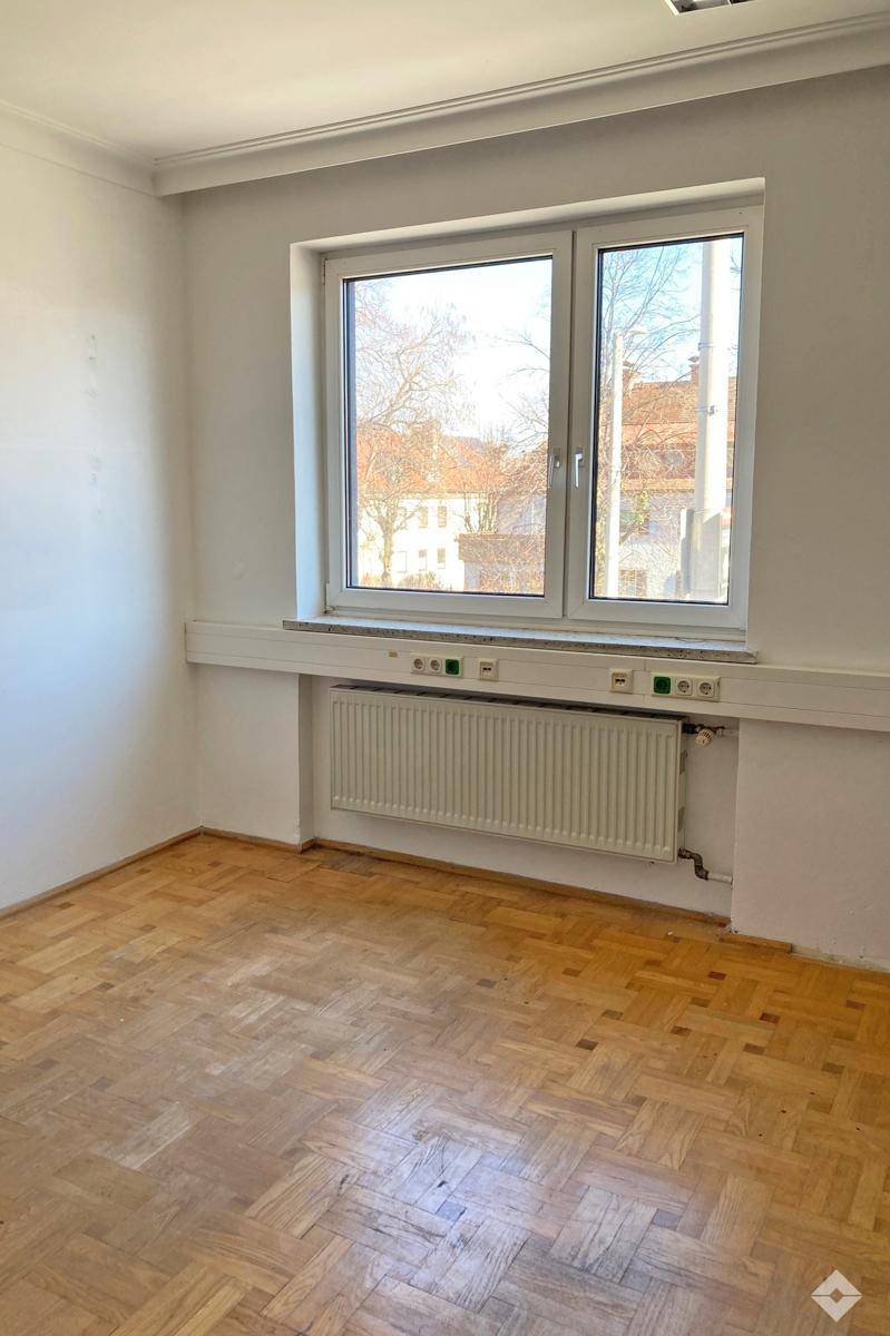 Leeres Büro mit blauem Teppichboden, weißen Wänden und einem Fenster mit Blick auf die Umgebung.