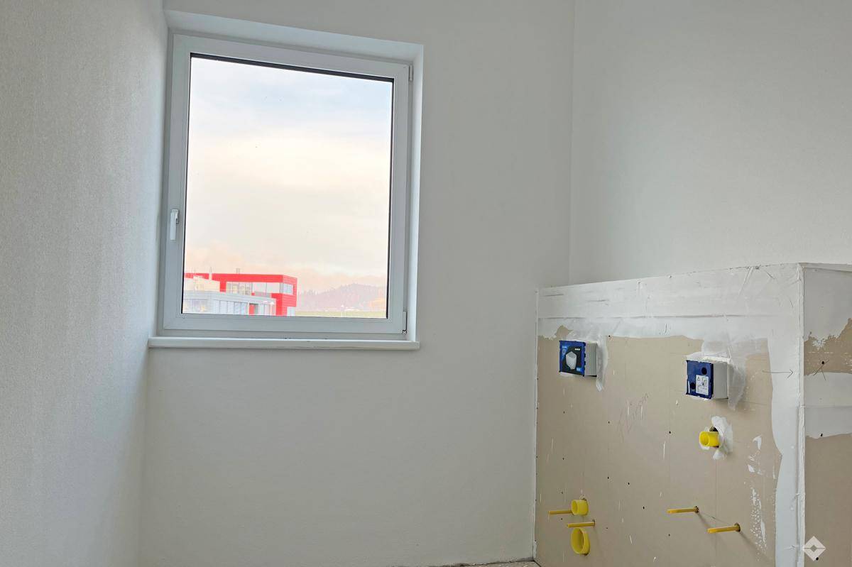 Unfertiges Badezimmer mit weiß gestrichenen Wänden, einem Fenster und vorbereiteten Anschlüssen für Sanitäranlagen.