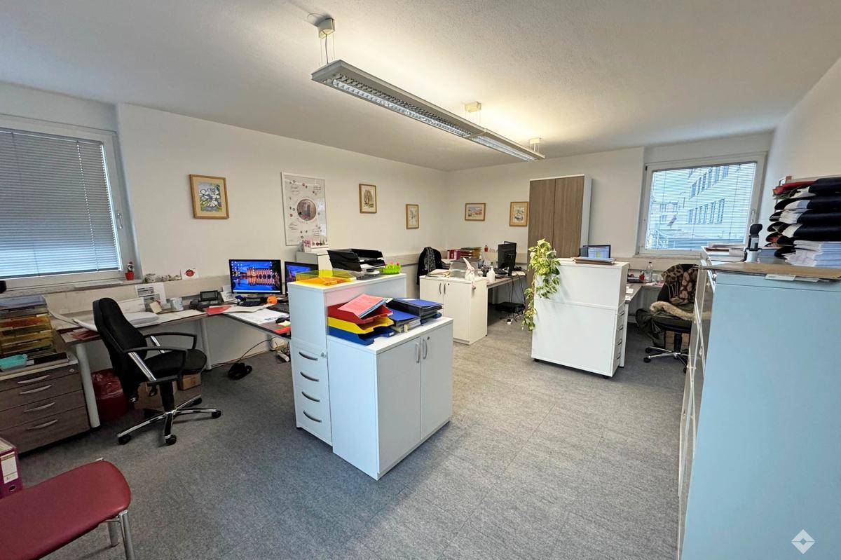 Großraumbüro mit mehreren Arbeitsplätzen, grauem Teppichboden und viel Tageslicht.