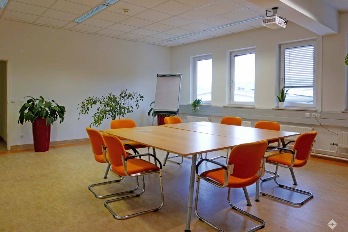 Großer Besprechungsraum mit rechteckigem Tisch, orangefarbenen Stühlen und Deckenprojektor.