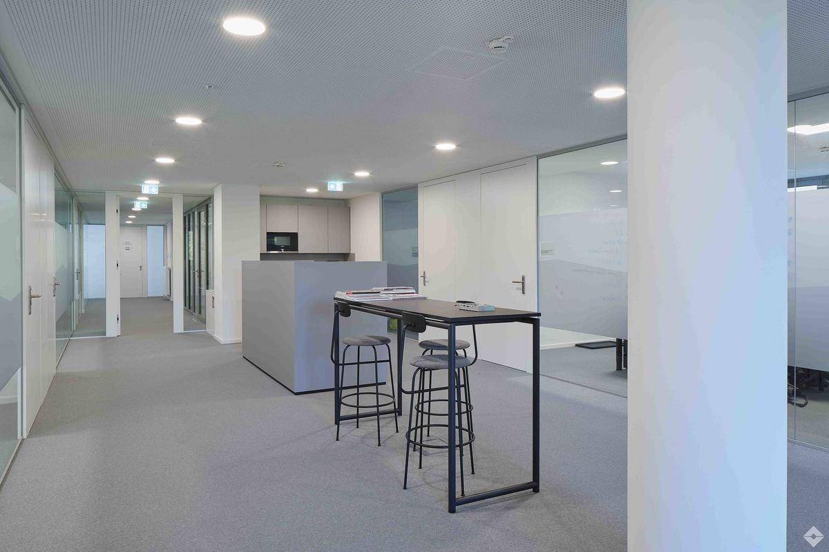 Moderner Bürogang mit Stehtisch, Barhockern und Glaswänden, im Hintergrund eine kleine Teeküche.