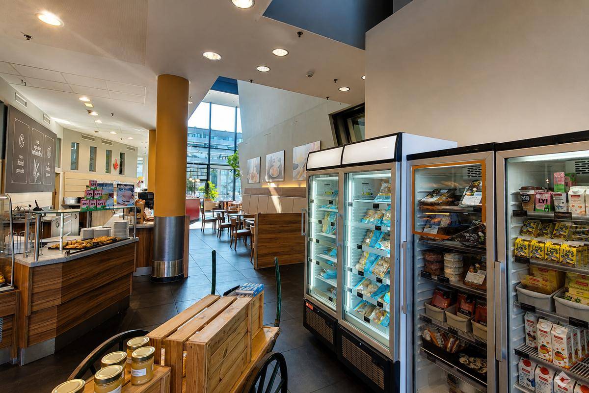 Verkaufsraum einer Bäckerei mit Kühlregalen, Holzkisten und Sitzbereich im Hintergrund.