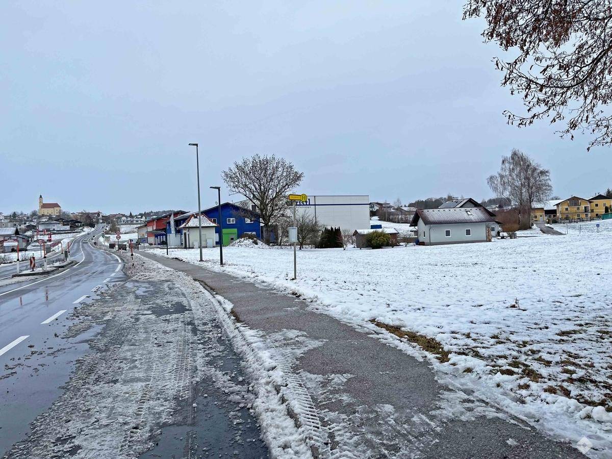 Blick auf ein schneebedecktes Feld neben einer Straße mit Gehweg und Gebäuden im Hintergrund.