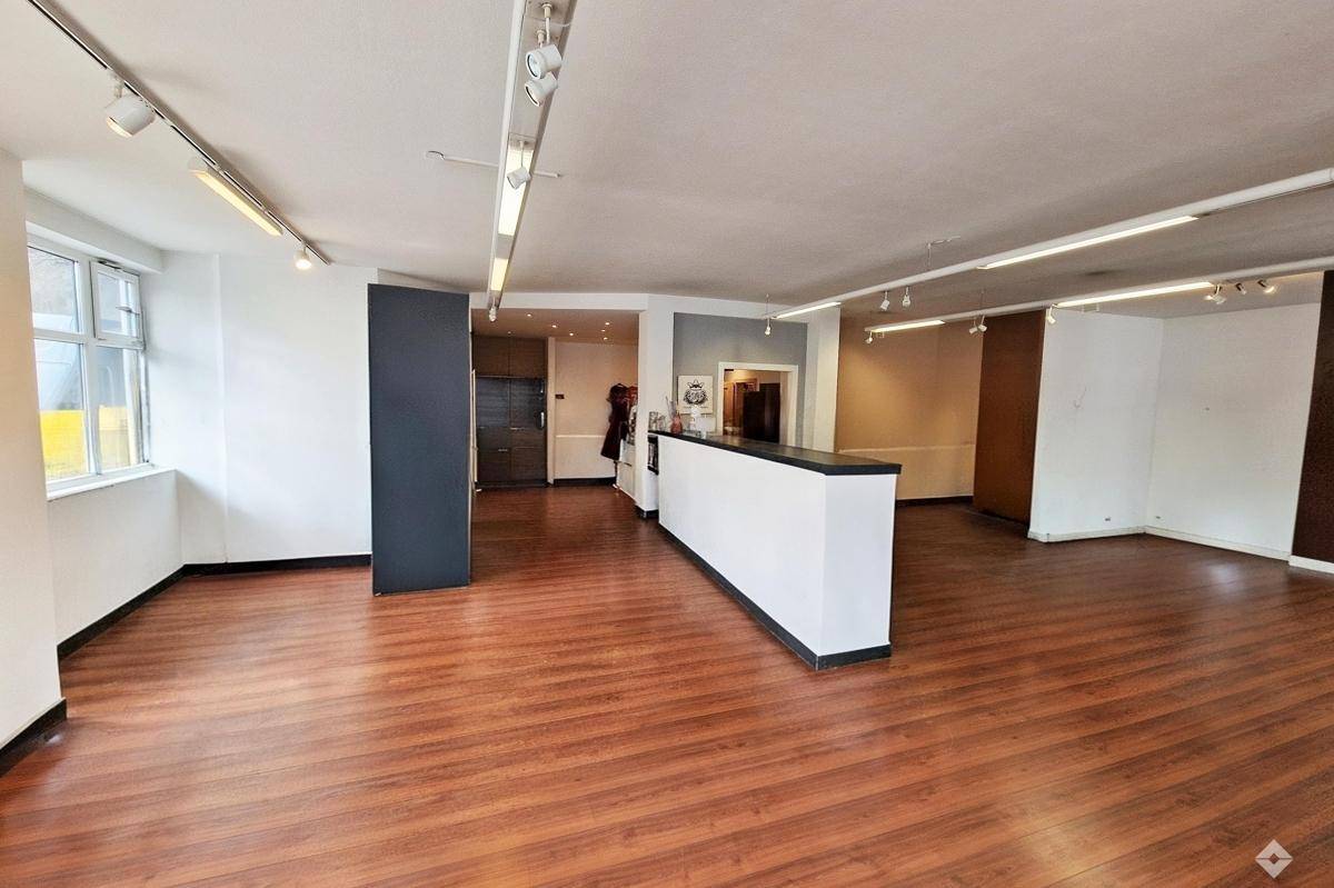Geräumiger, leerer Geschäftsraum mit Holzboden, Theke und Schienenbeleuchtung an der Decke.