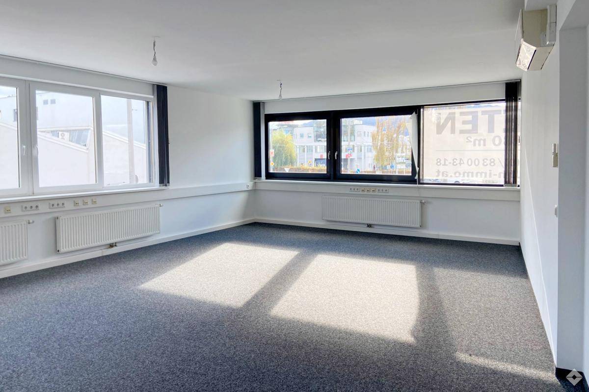 Heller Büroraum mit Teppichboden, großen Fenstern und Blick auf die Stadtumgebung.