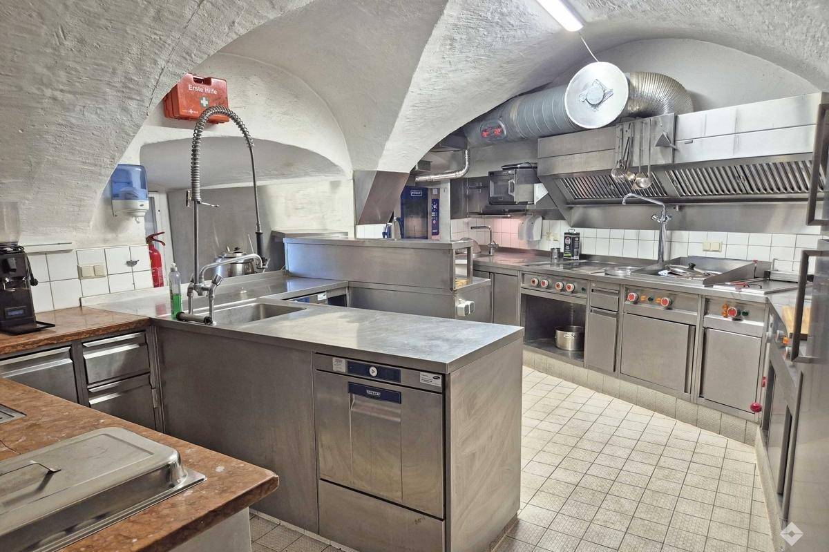 Professionelle Edelstahlküche mit Fliesenboden, Dunstabzugshaube und gewölbter Decke.