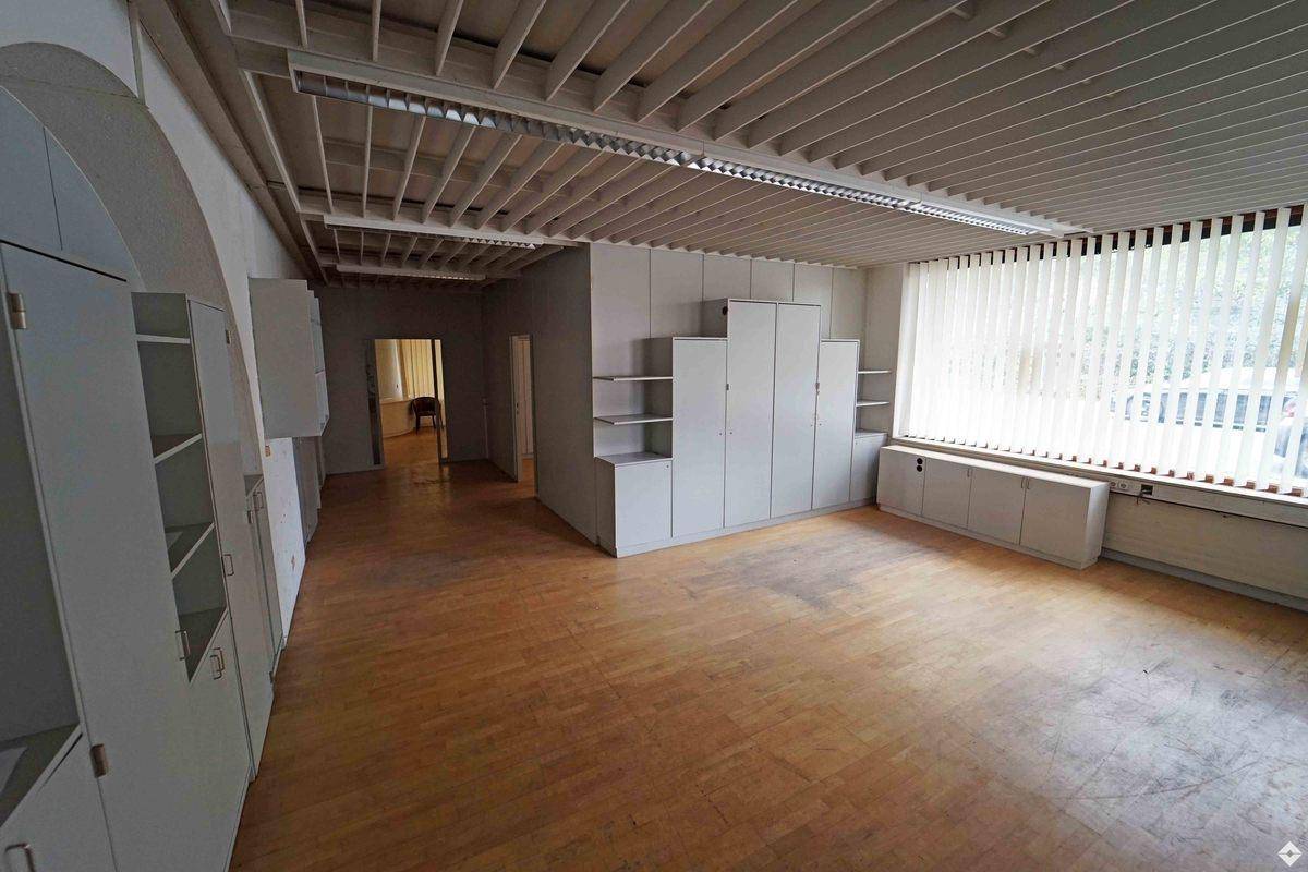 Geräumiger Büroraum mit Holzparkettboden, Einbauschränken und großen Fenstern mit Jalousien.