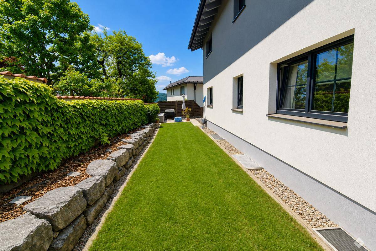 Gepflegter Gartenbereich mit Rasenfläche, Hecke und Steinmauer entlang des Hauses.