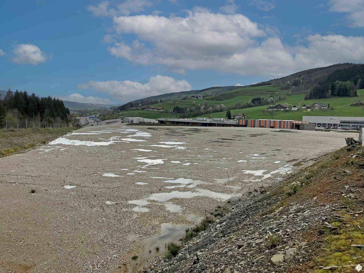 Großes, unbebautes Kiesgrundstück mit Wasseransammlungen, im Hintergrund grüne Hügel und Gewerbebauten bei sonnigem Wetter.