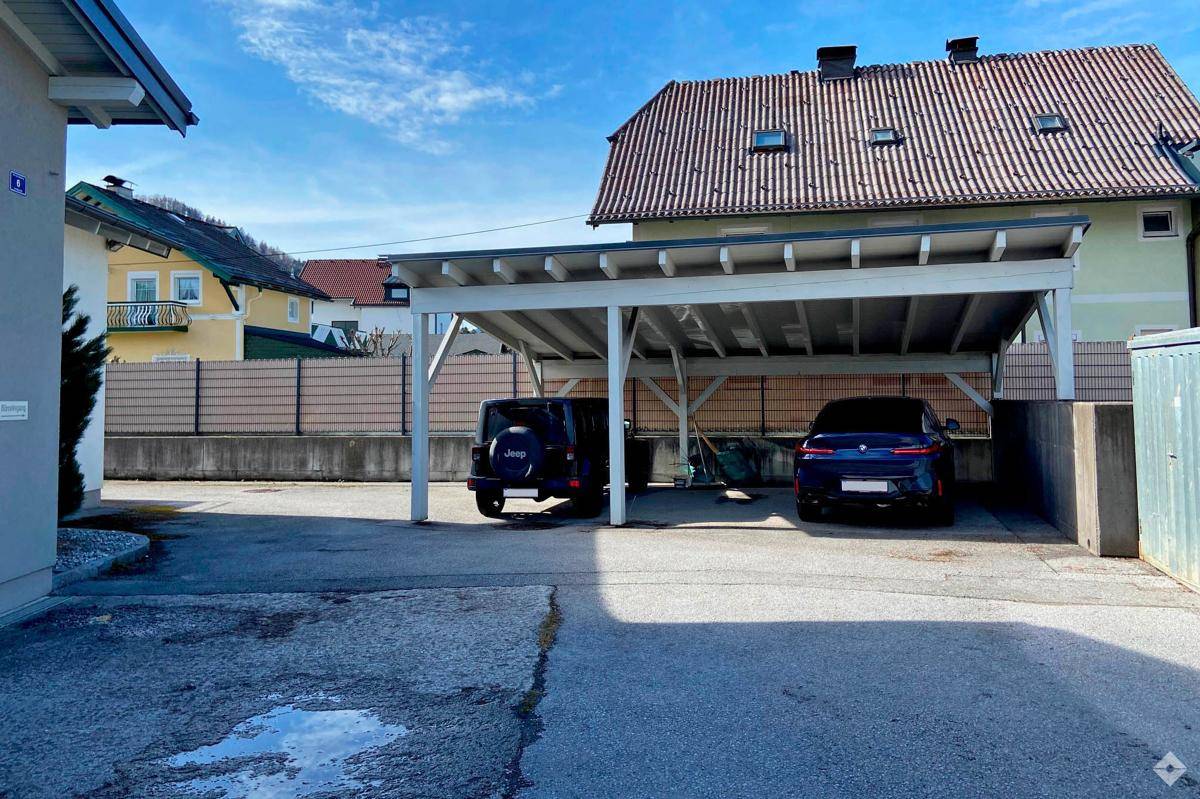 Geräumiger Carport mit zwei geparkten Autos und einem angrenzenden Gebäude unter blauem Himmel.