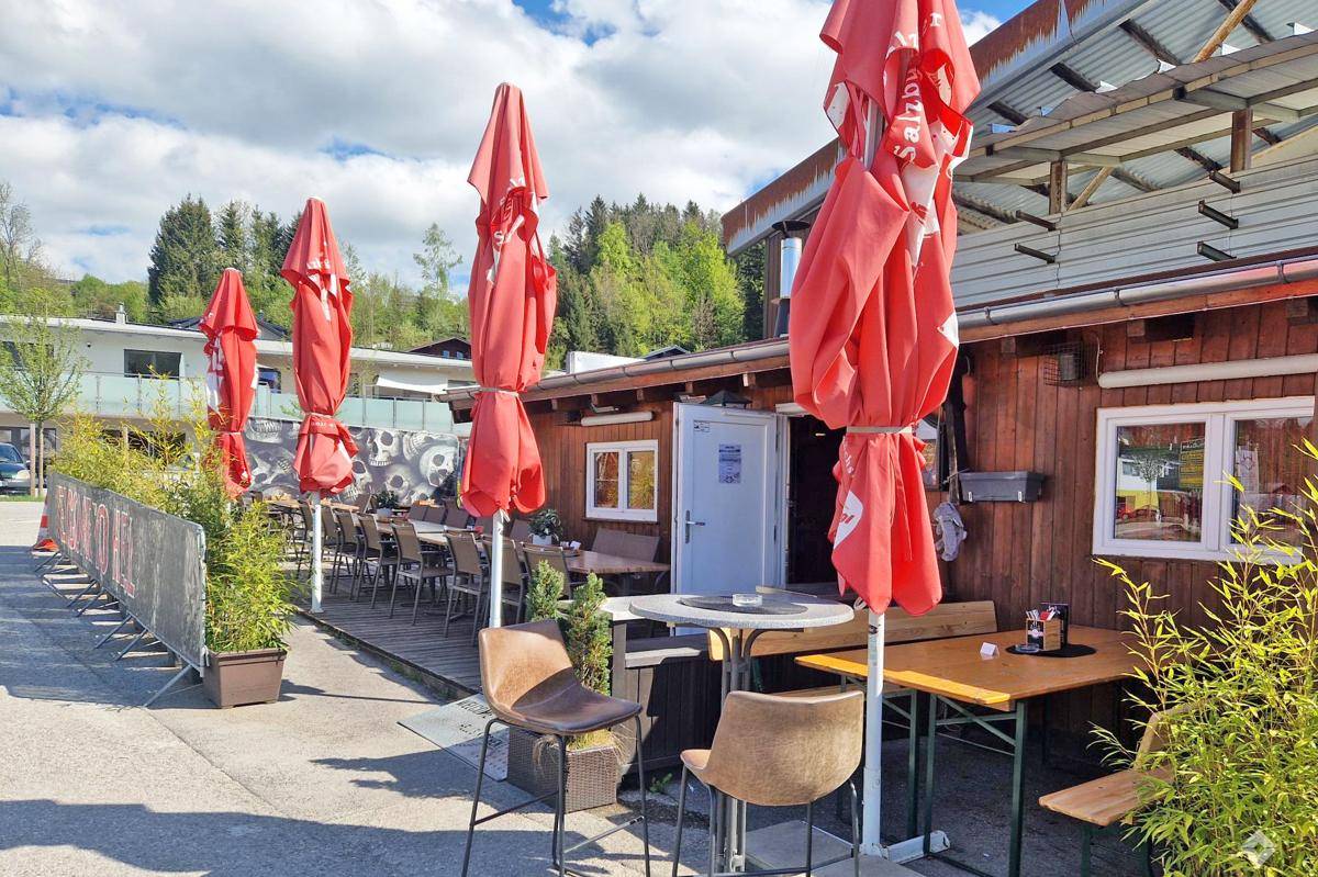 Eingangsbereich des Holzgebäudes mit weiteren Sitzgelegenheiten im Freien und hohen Tischen.