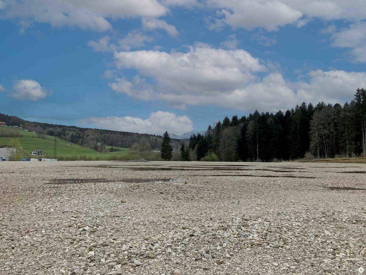 Ausgedehntes Kiesgrundstück mit Waldrand und grünen Hügeln im Hintergrund, unter einem weiten blauen Himmel.