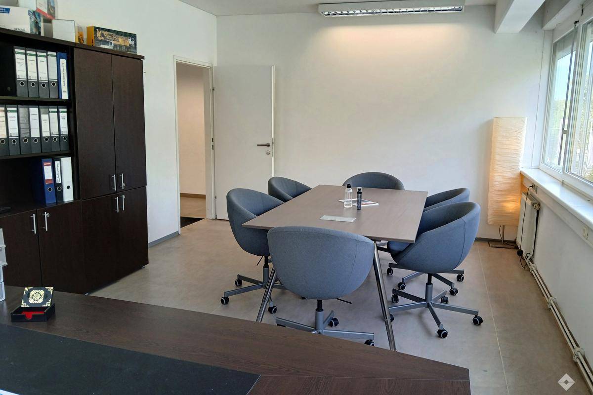 Heller Büroraum mit Besprechungstisch, modernen Stühlen und großem Fenster mit Jalousien.