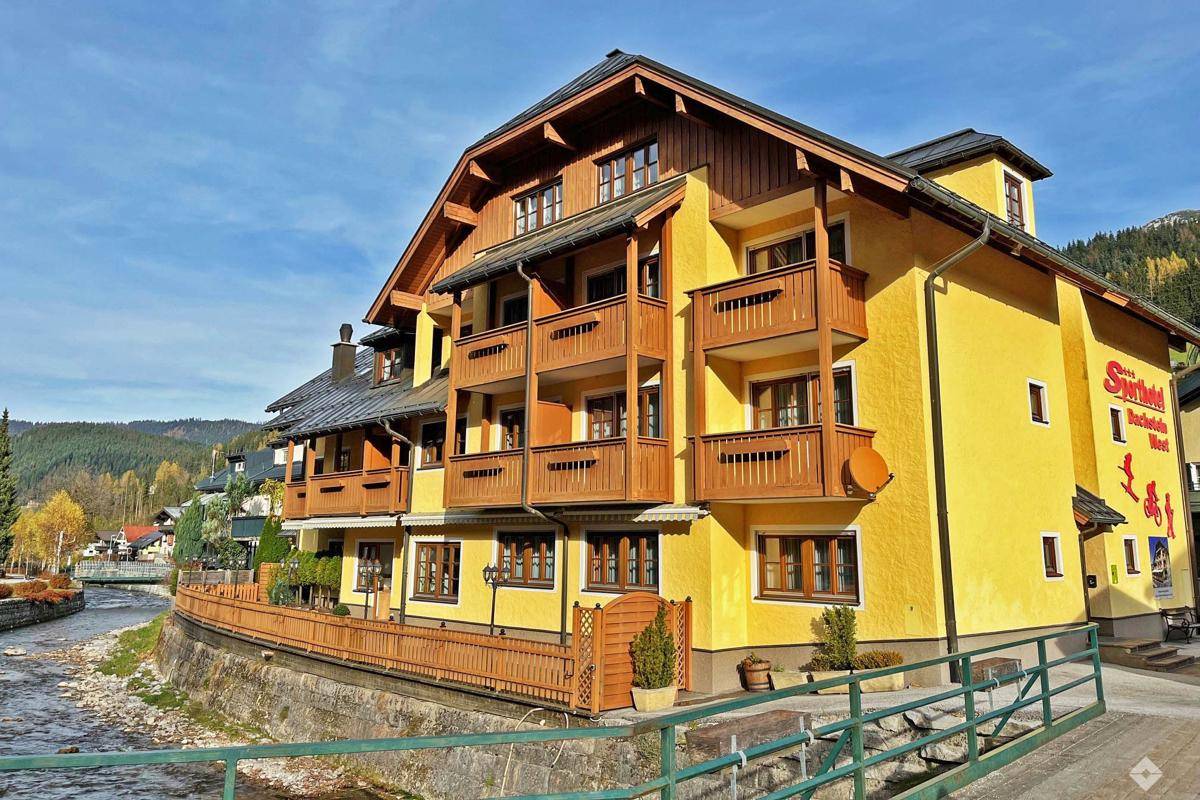 Gelbes Gebäude mit Holzbalkonen und traditioneller Architektur am Flussufer, umgeben von Bergen.